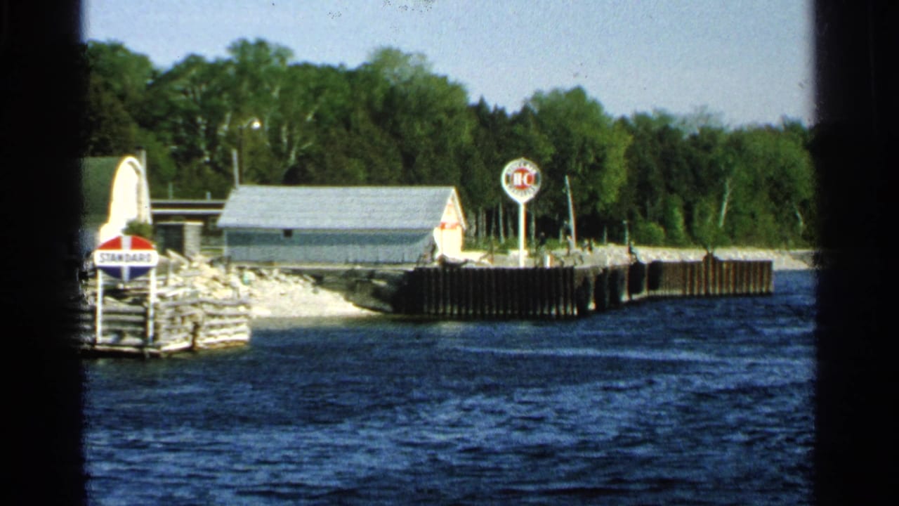 1964 Washington Island lakeside scene with a small building, pier wall, Standard sign, circular HC sign, trees, and rippled water.