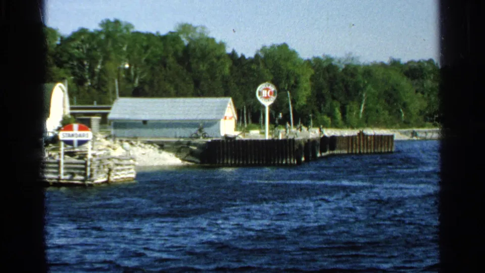 Washington Island dock area in 1964, showing the Standard sign, small shore building, wooden pier, rocks, trees, and dark blue water.