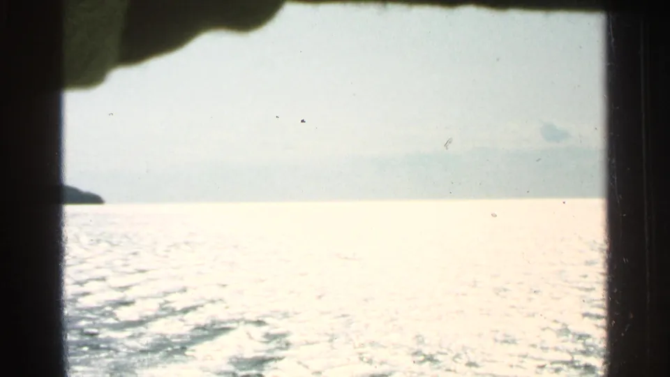 Bright overexposed lake view from the 1964 Washington Island footage, with pale sky, reflective water, and a dark obstruction near the frame edge.