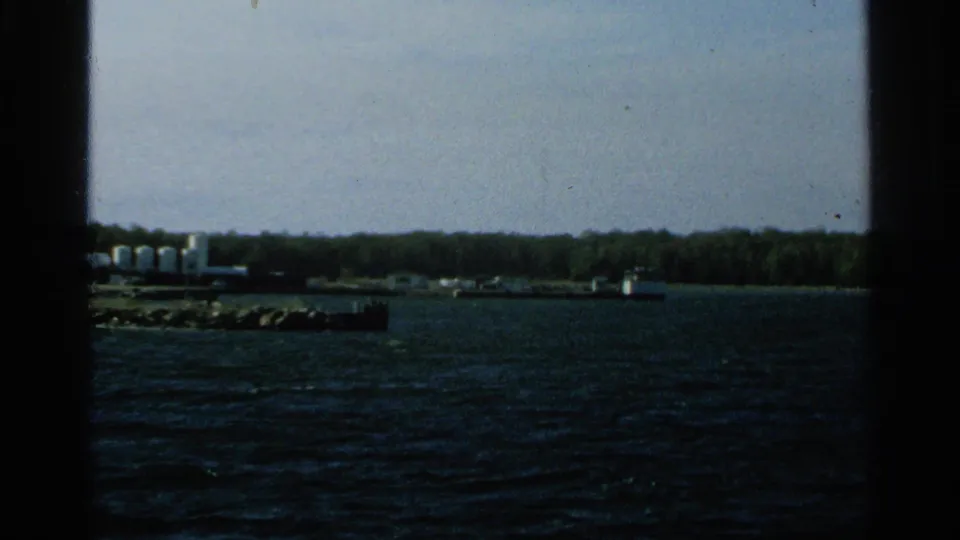 1964 Washington Island shoreline view with tree cover, low waterfront structures, and choppy dark water in the foreground.