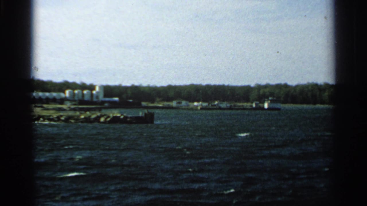 1964 Washington Island shoreline view from a boat, with dark water, white storage tanks, low buildings or dock structures, and trees.