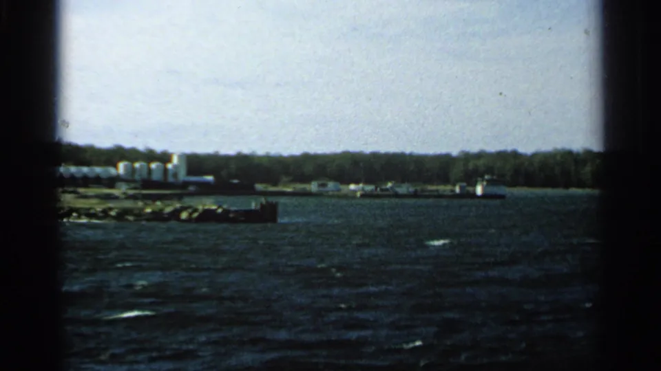 1964 Washington Island shoreline view from a boat, with dark water, white storage tanks, low buildings or dock structures, and trees.