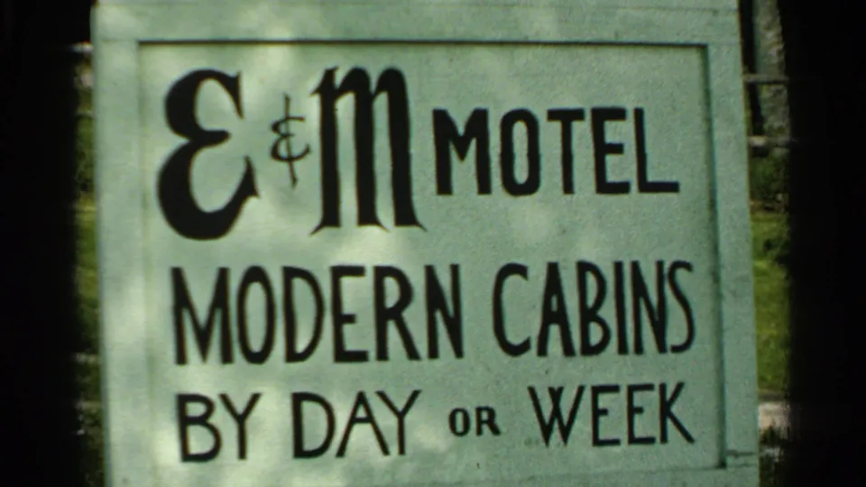 1964 Washington Island frame showing an E&M Motel sign reading modern cabins by day or week, partly framed by shadow and greenery.