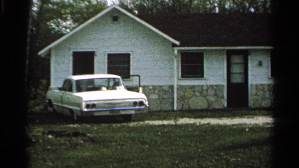 Motel cabin exterior on Washington Island in 1964, showing the white building, stone base, parked sedan, grass, and surrounding trees.