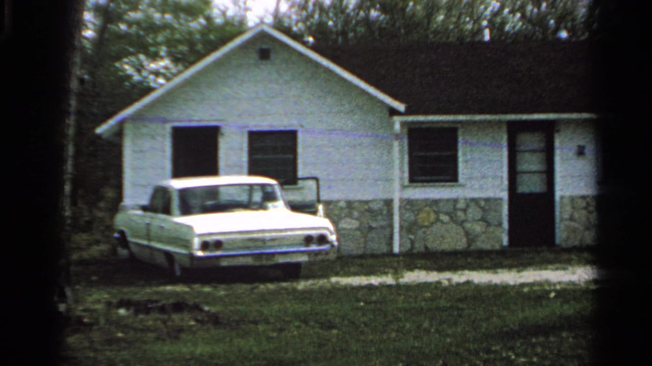 White car parked in front of a small white house with a stone foundation.