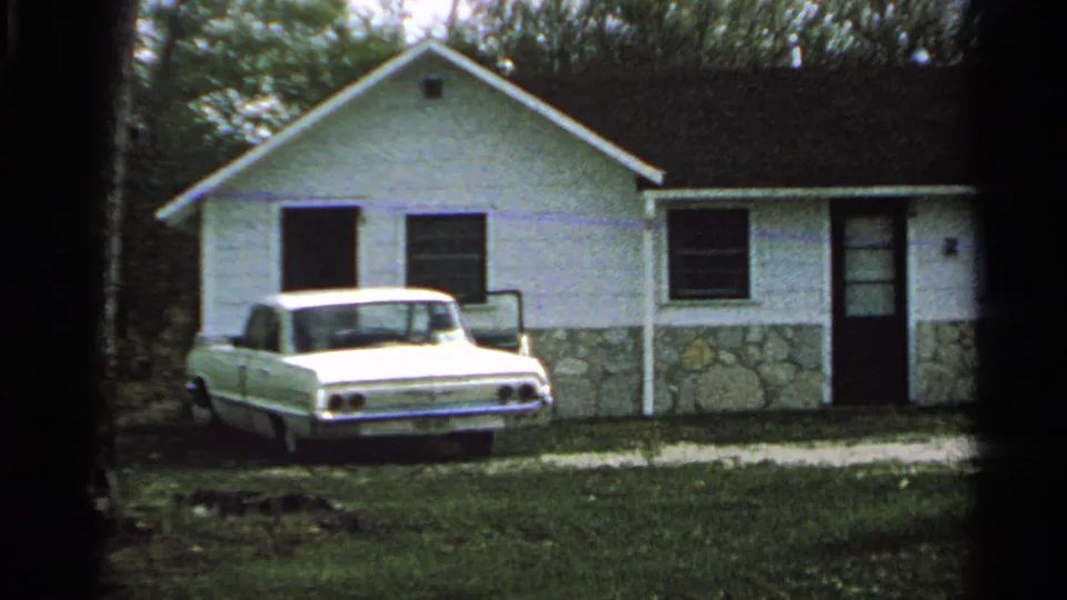 1964 Washington Island home-movie frame showing a white car outside a modest white house with trees and grass around it.