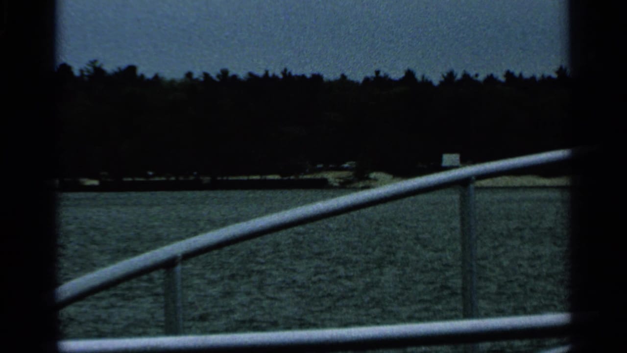 1964 Washington Island 8mm frame showing a boat railing, lake water, dark film edges, and a distant tree-lined shore.