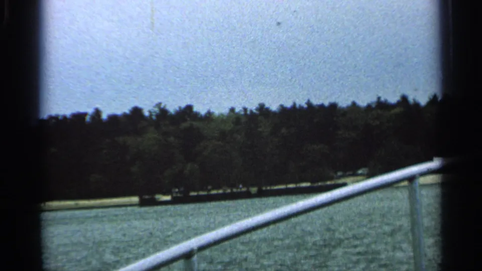 1964 8mm frame from Washington Island showing a moving boat perspective toward water, trees, and a pale shoreline strip.