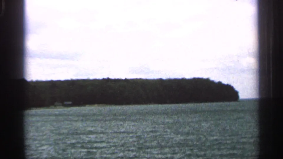 1964 Washington Island shoreline view with open lake water, a wooded point, bright sky, and visible 8mm frame edges.