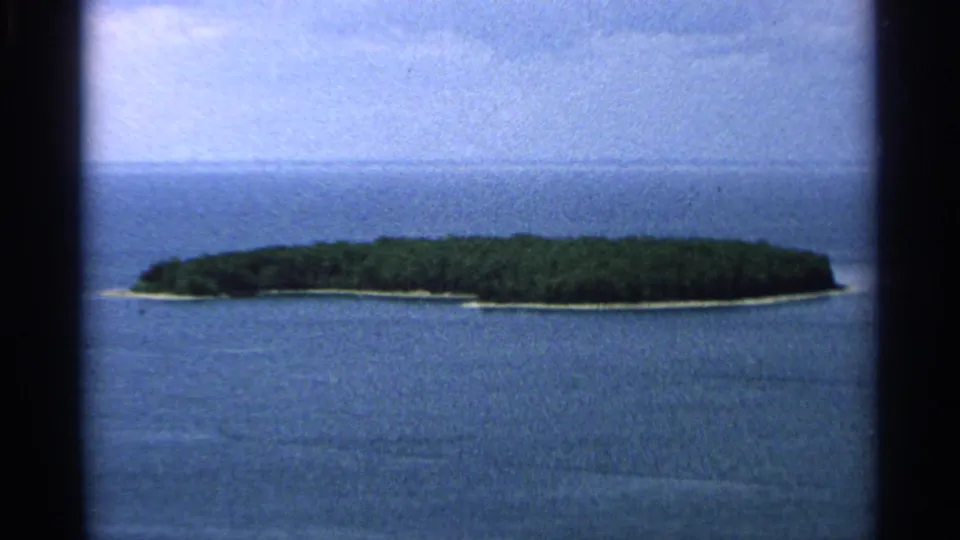1964 Wisconsin 8mm frame showing a low green island encircled by calm blue water under a pale sky.