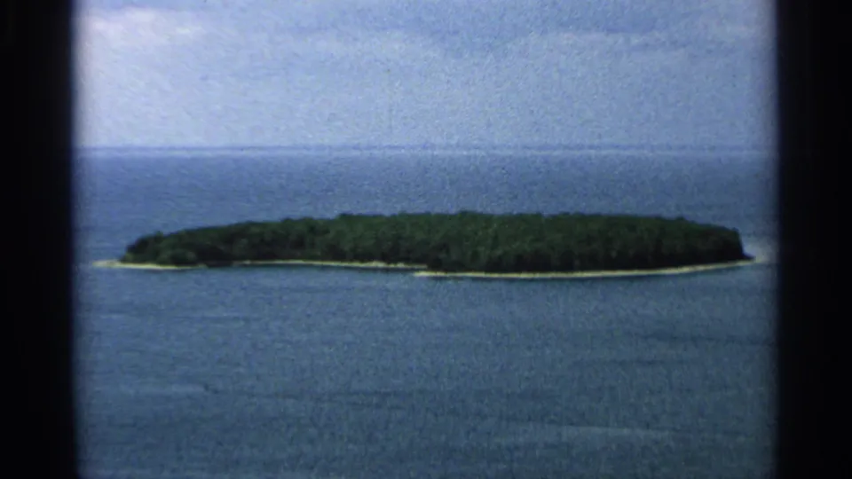 1964 Wisconsin home-movie frame showing a wooded island, narrow shoreline, blue water, and soft sky.