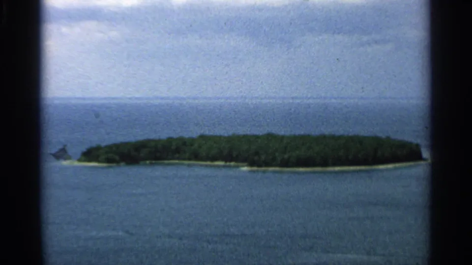 1964 8mm aerial view of Washington Island, Wisconsin, with a low wooded shoreline, surrounding blue water, and a distant horizon.