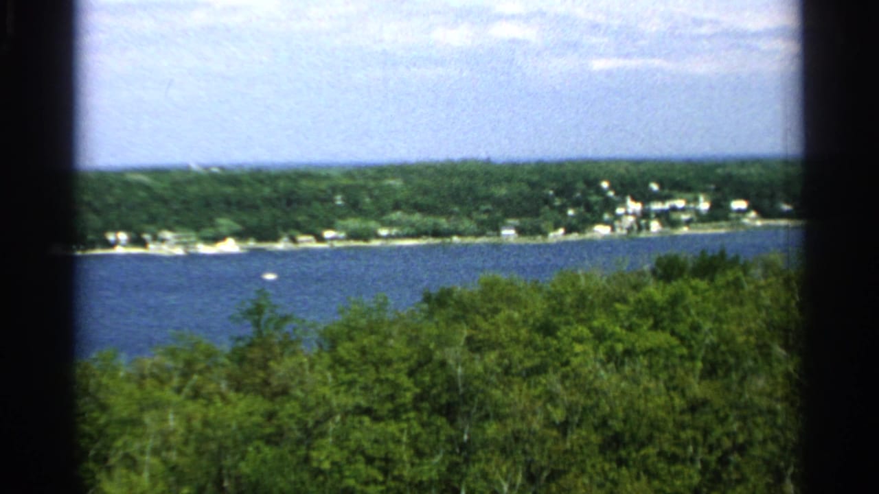 1964 Washington Island, Wisconsin home-movie view over blue water toward a wooded shoreline with scattered light buildings.