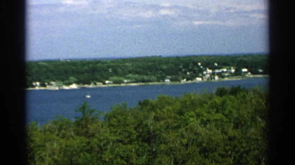 1964 Washington Island, Wisconsin shoreline with blue water, dense green trees, and small distant buildings near the shore.