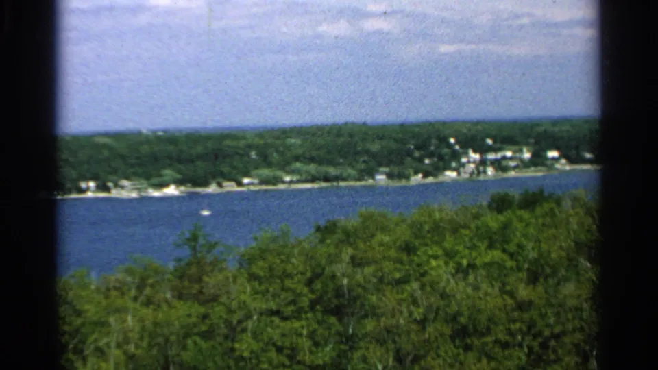 1964 Washington Island, Wisconsin view of a small boat crossing blue water below a wooded shore and scattered houses.