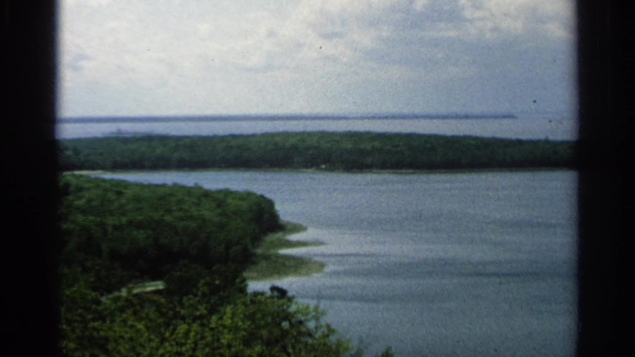Washington Island shoreline in 1964, with blue-green water, tree-covered land, cloudy sky, and dark structural edges in front.