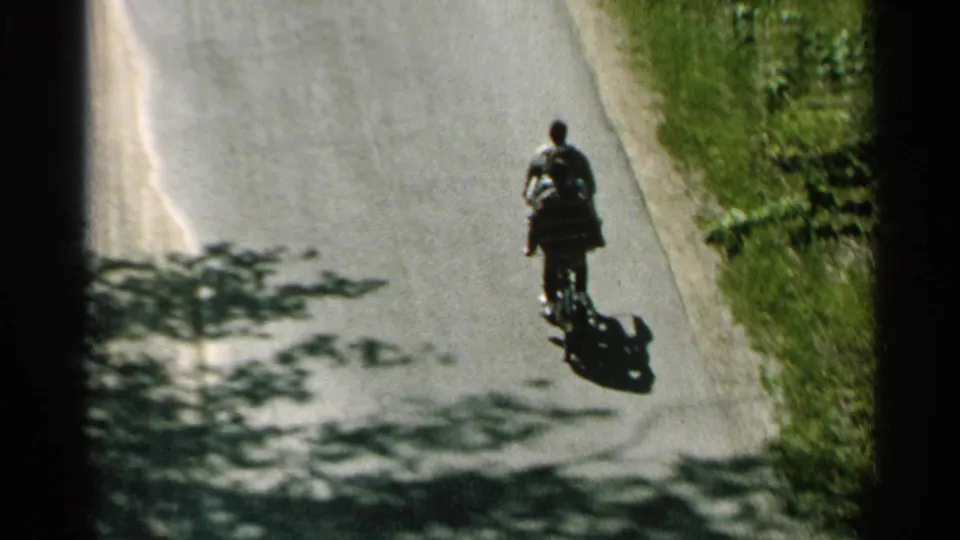 In 1964 Washington Island footage, two distant riders share a bicycle on a paved road bordered by grass and tree shadows.