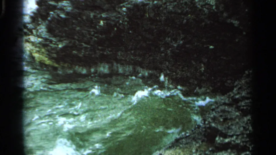 1964 Washington Island 8mm frame showing rough green water and white spray against dark, mossy rock surfaces.