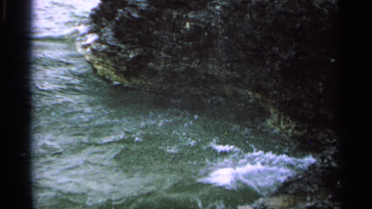 Green-tinted 8mm view of water surging between dark Washington Island rocks, with small whitecaps and foam near shore.