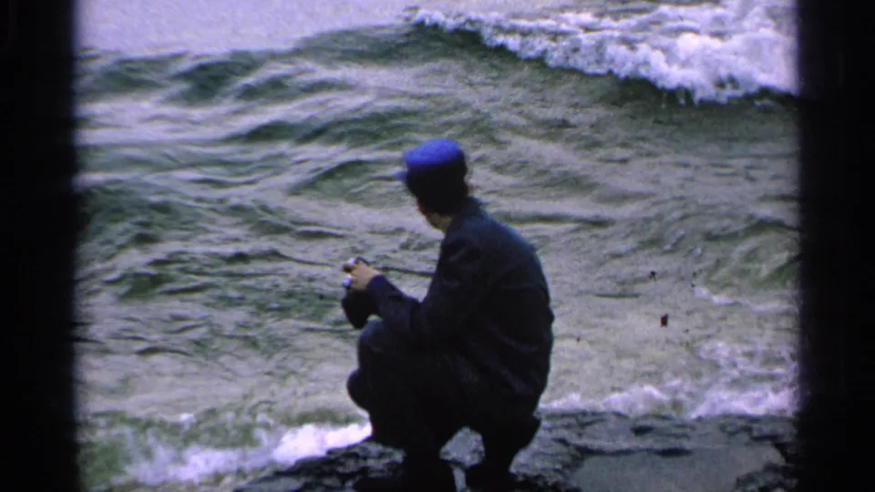 A man in a dark jacket and blue cap crouches on Washington Island rocks, facing waves and foam near the shore.