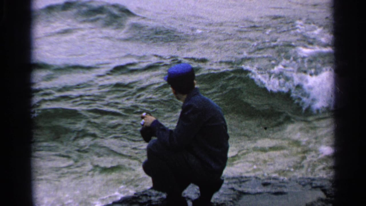 Man in a blue cap crouches on rocks beside churning water.