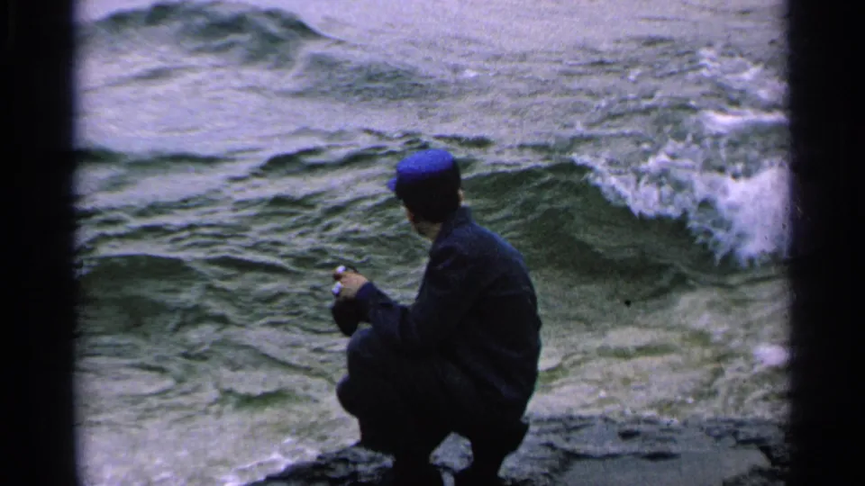 In 1964 Washington Island home-movie footage, a man in a blue cap crouches on a rocky shoreline near foaming waves.
