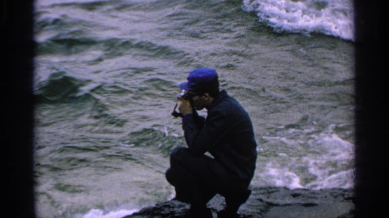 The man in a blue cap holds a small movie camera to his face while crouched beside rough shoreline water.