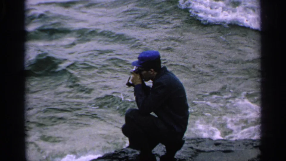 The man in a blue cap holds a small movie camera to his face while crouched beside rough shoreline water.