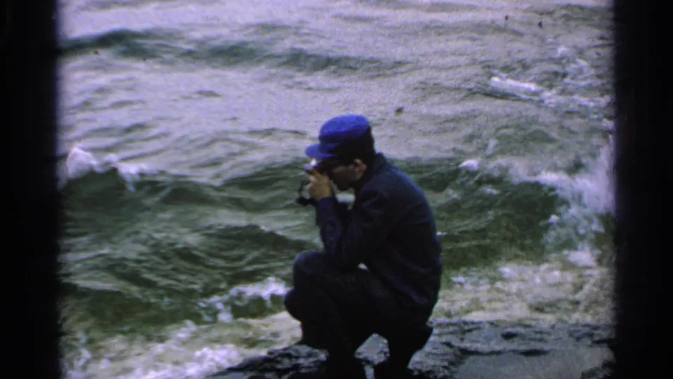 A handheld 8mm frame shows the crouching cameraman facing the water as waves break below the rocks.