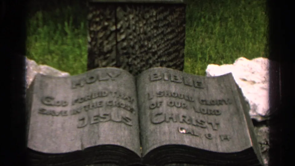 A stone sculpture shaped like an open book displays raised religious lettering beside grass and a dark vertical stone surface.