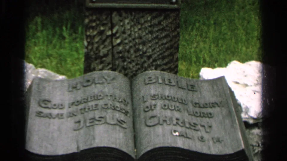 Washington Island 1964 home-movie frame showing the open-book grave marker with visible HOLY BIBLE text and surrounding grass.