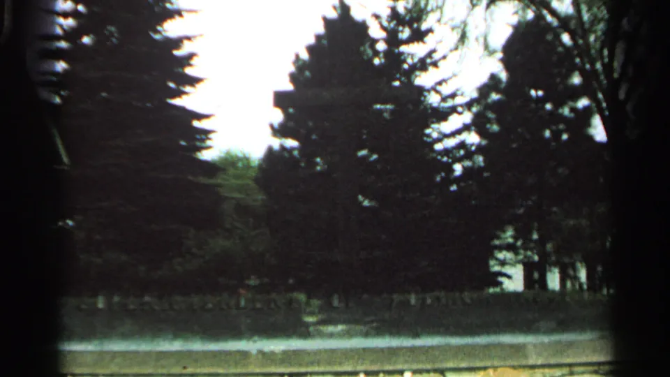 The camera drops toward the Nicolet Memorial area, showing trees and hedge above the partially visible plaque edge in 1964 home-movie footage.