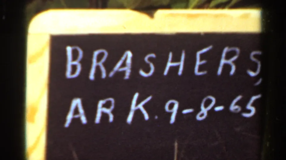 Tight 1965 Arkansas home-movie frame showing the Brashers chalkboard date slate with soft focus and visible film grain.