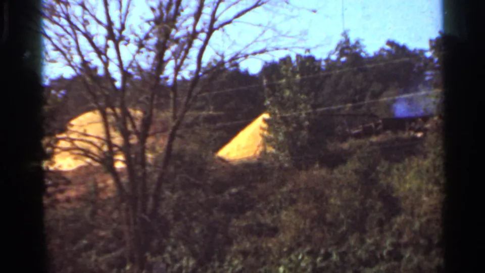 1965 Arkansas home-movie frame with brush, bare trees, utility lines, and distant yellow-roofed structures in soft focus.