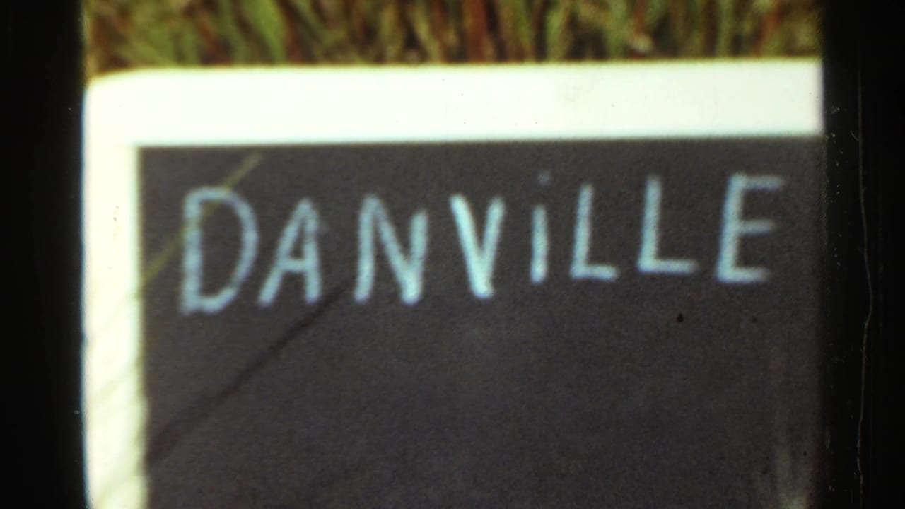 1965 Arkansas home-movie frame showing a close view of a hand-painted Danville sign bordered in white with grass behind it.
