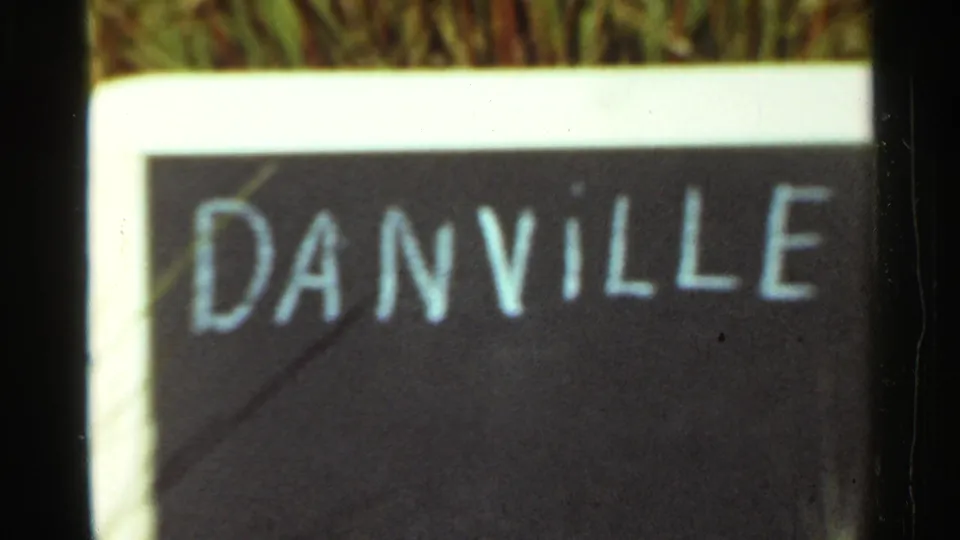 1965 Arkansas home-movie frame showing a close view of a hand-painted Danville sign bordered in white with grass behind it.