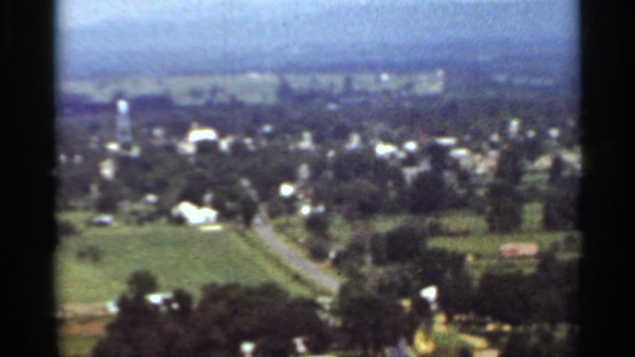 1965 Arkansas 8mm view over a small town, with scattered houses, green fields, roads, trees, and a hazy horizon.