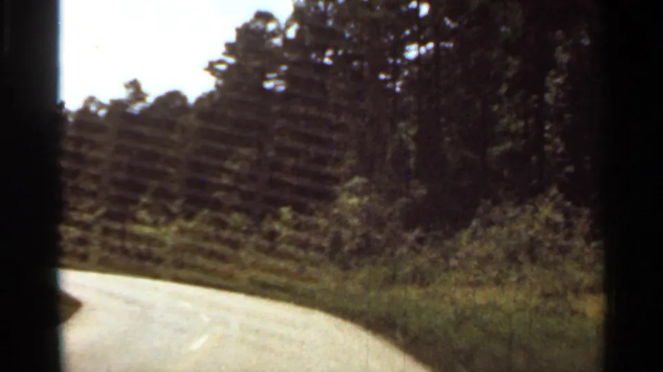 Passenger-view 8mm frame of a road bending through a wooded Arkansas area, with trees and brush lining the shoulder.