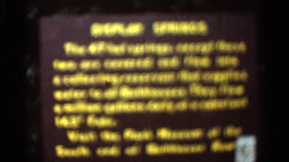 Arkansas 1965 home-movie frame showing a brown park display sign with yellow lettering about springs, partly blurred.