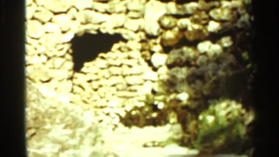 1965 Arkansas home-movie view of a rough stone cave-like wall with a dark arched opening and dry vegetation.