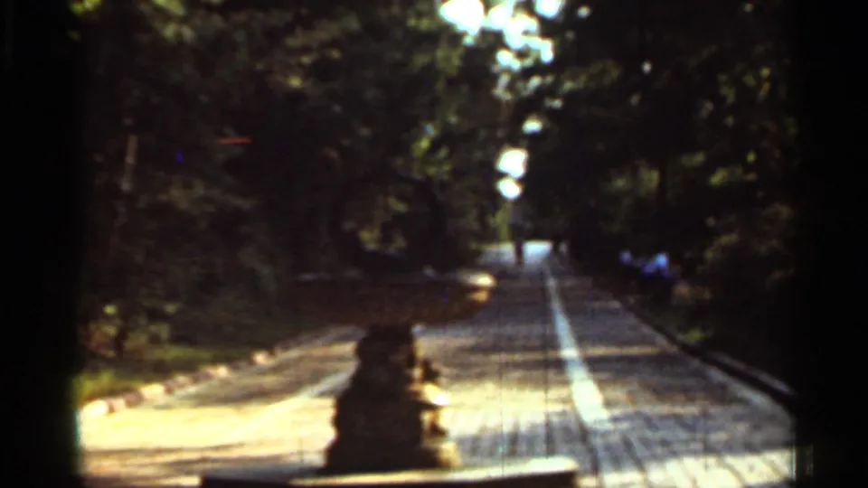 1965 Arkansas 8mm frame of a park walkway, foreground stone fountain or post, leafy shade, and indistinct people in the distance.