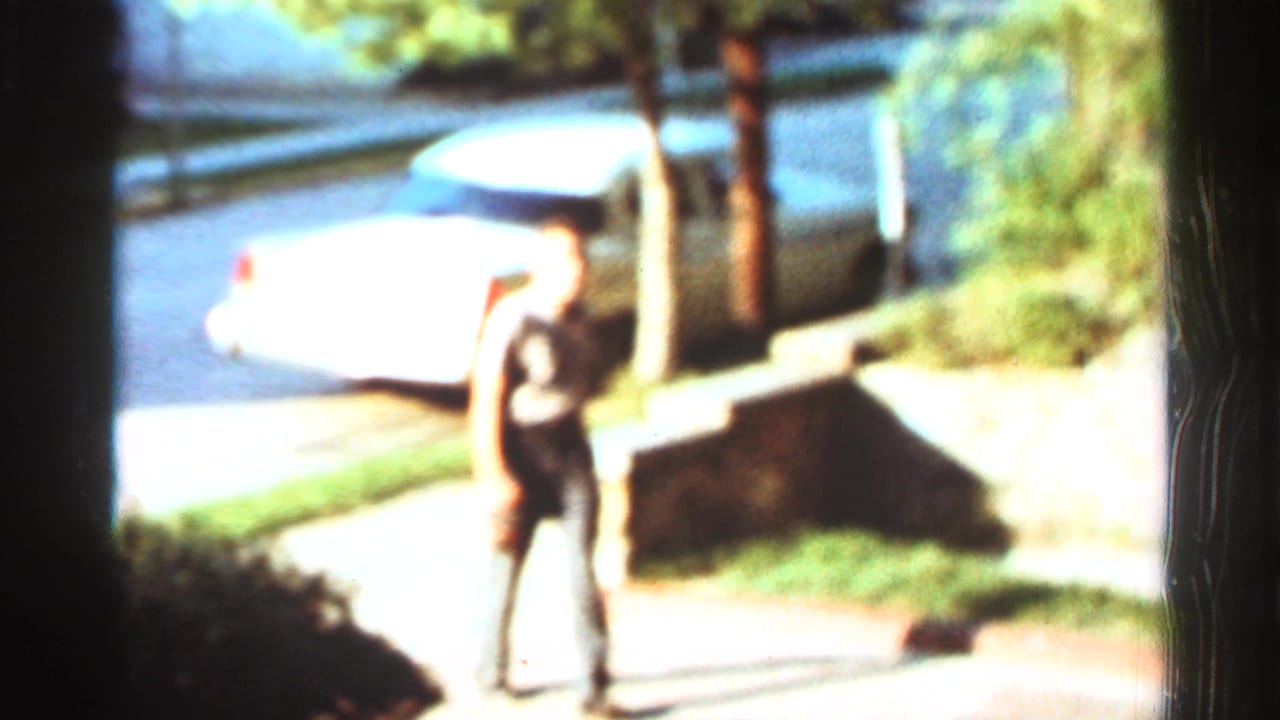 Adult man walking on a sidewalk near a white parked sedan and stone wall.