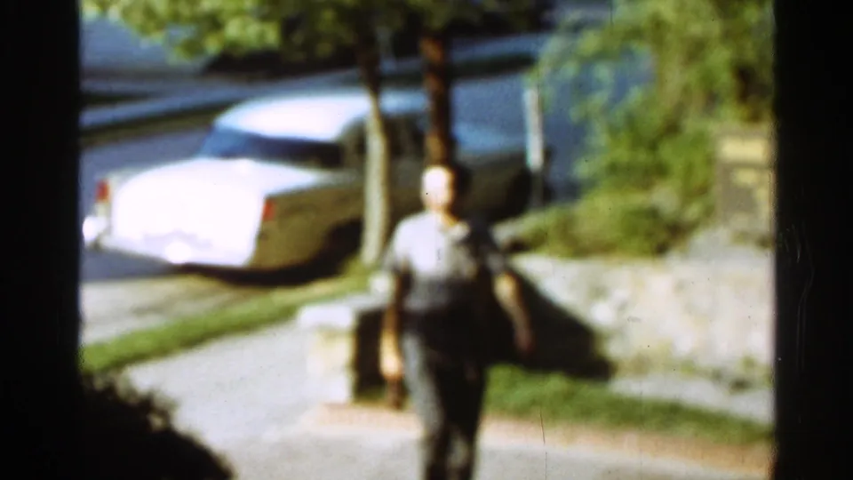 Soft 8mm frame from Arkansas in 1965 showing a man walking toward the camera with a white sedan and street behind him.