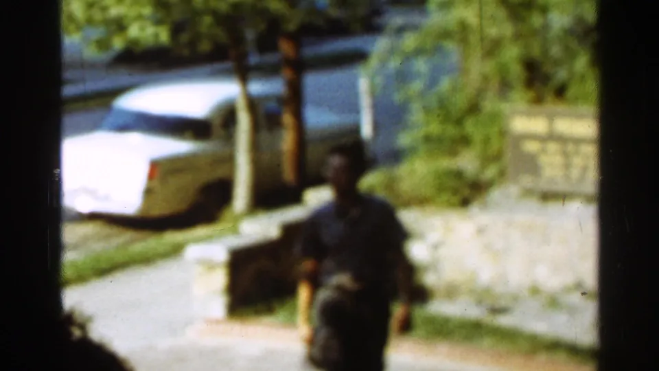 1965 Arkansas 8mm frame of the man seen from behind as he continues along the sidewalk beside the parked white sedan.