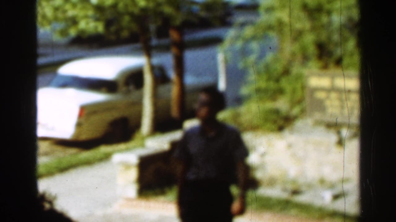 1965 Arkansas home-movie frame showing a man walking along a sidewalk near a white 1960s sedan on a leafy street.