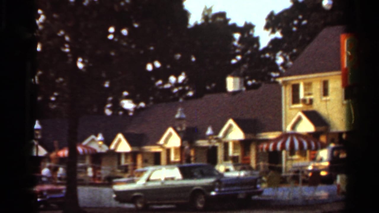 1965 Arkansas home-movie view of small roadside amusement shops with striped awnings, trees, and a classic car in front.