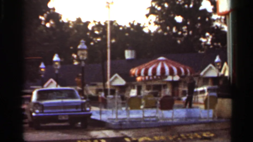 1965 Arkansas amusement-area view with a striped round canopy, classic car, low fencing, decorative lamps, and distant visitors.