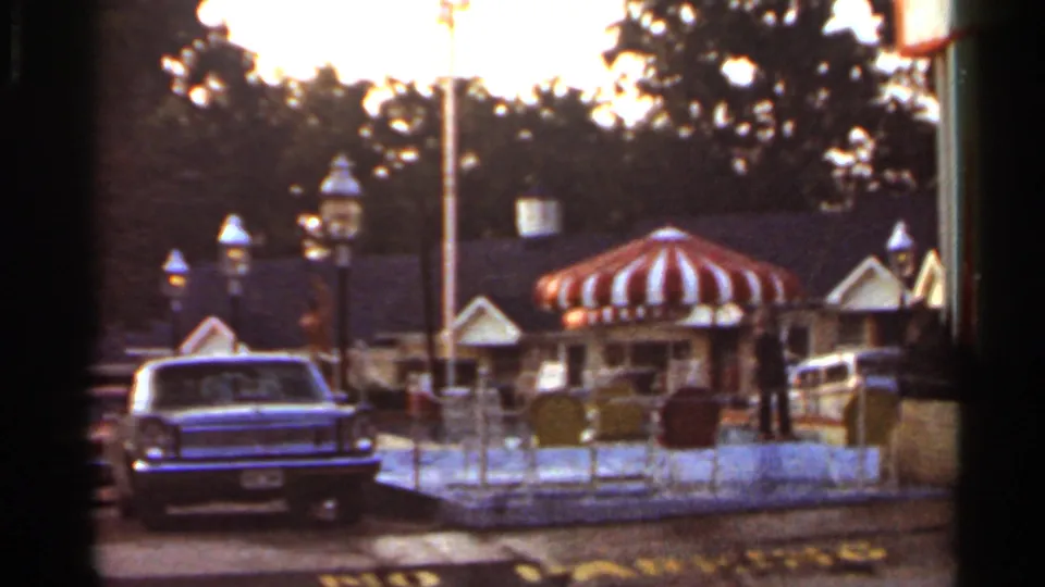 1965 Arkansas home-movie frame showing a striped canopy, vintage lamp posts, a parked classic car, and distant visitors near shop roofs.