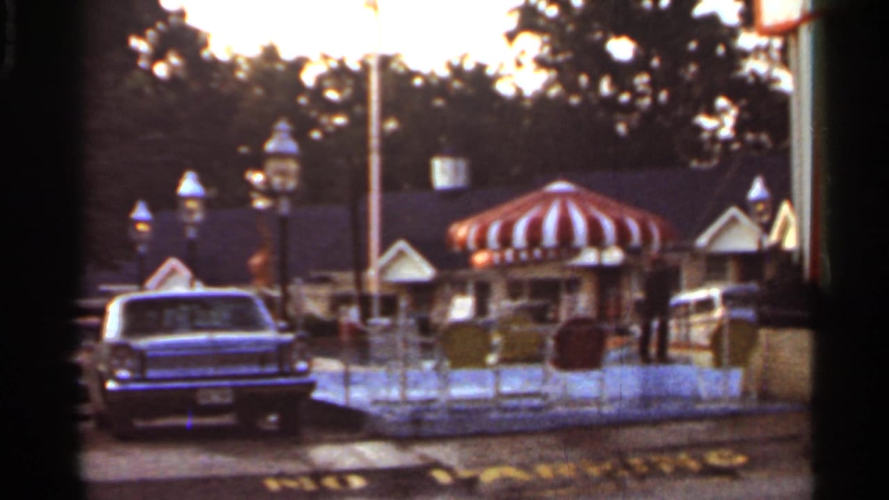 Motel exterior at dusk with a striped awning, parked sedan, lamps, and roadside fencing.