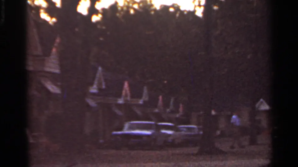 1965 Arkansas motel scene at dusk with parked vintage cars, peaked roof details, trees, and a person walking in the distance.